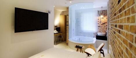 Habitación de hotel moderna con televisión de pantalla plana, una bañera grande, pared de ladrillo y un pequeño escritorio con silla.