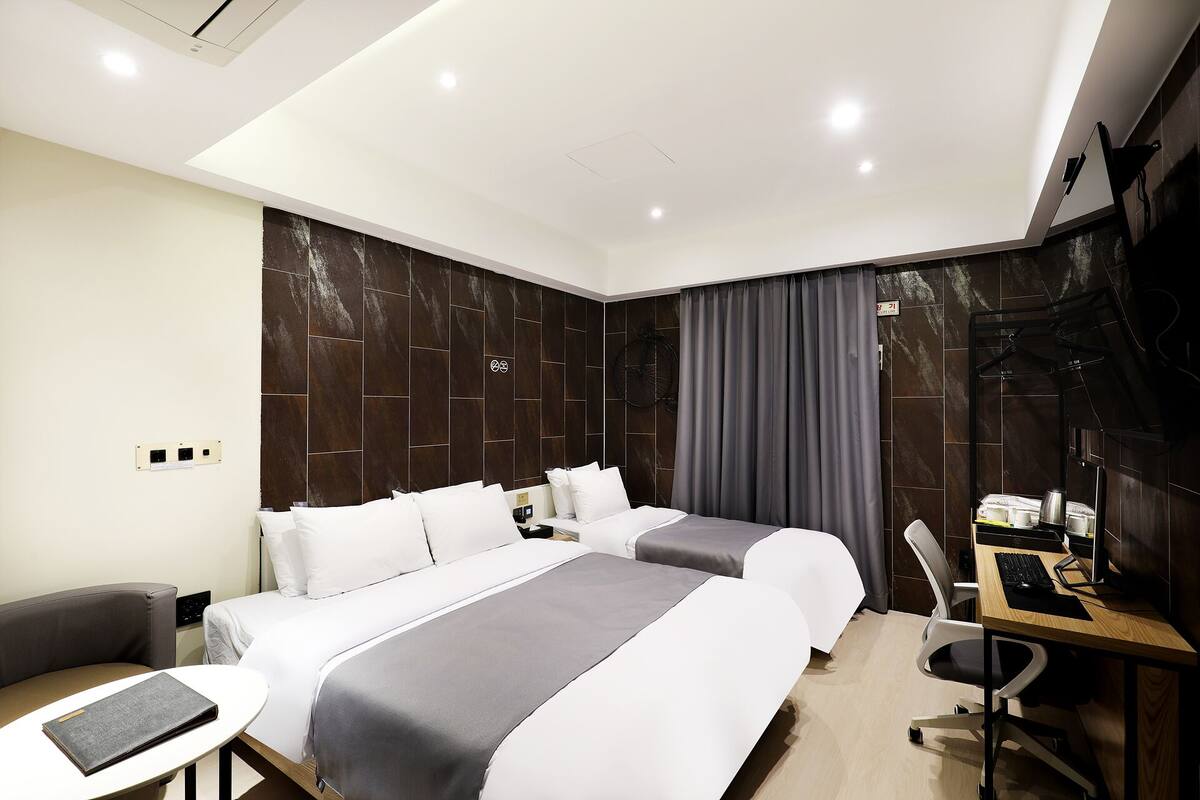 A hotel room with two beds, a desk with a computer, a chair, and a small table with a book.