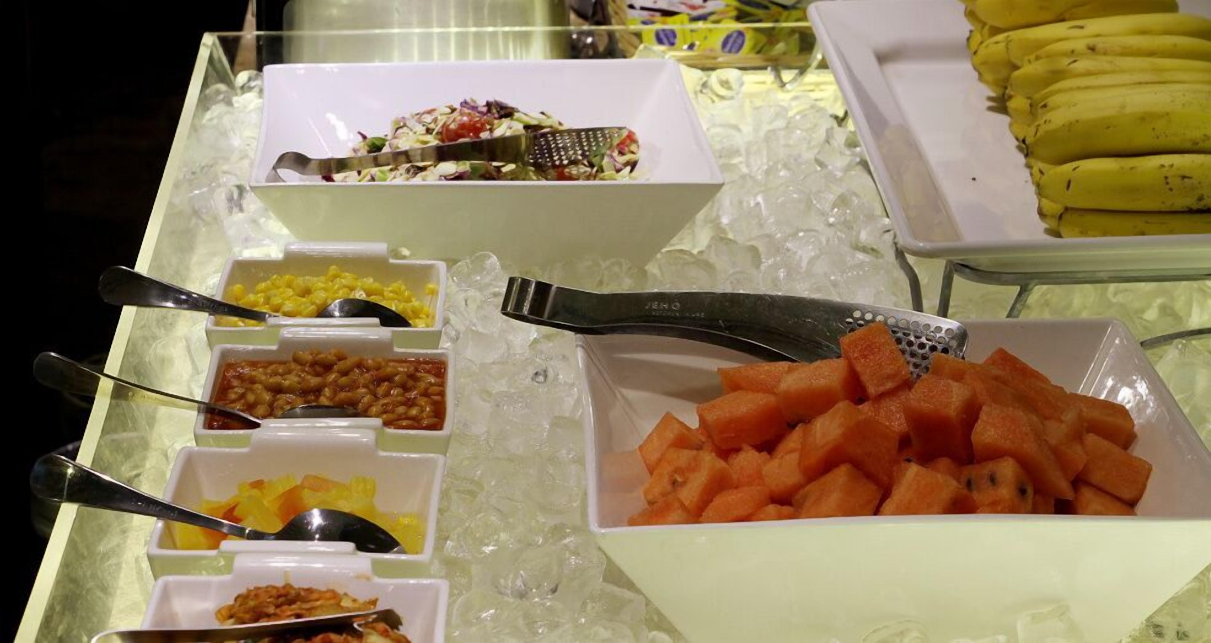Buffet com diversos alimentos, incluindo milho, feijão, salada e fruta em cubos.
