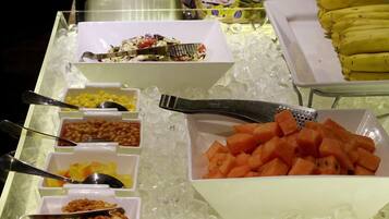 Buffet com diversos alimentos, incluindo milho, feijão, salada e fruta em cubos.