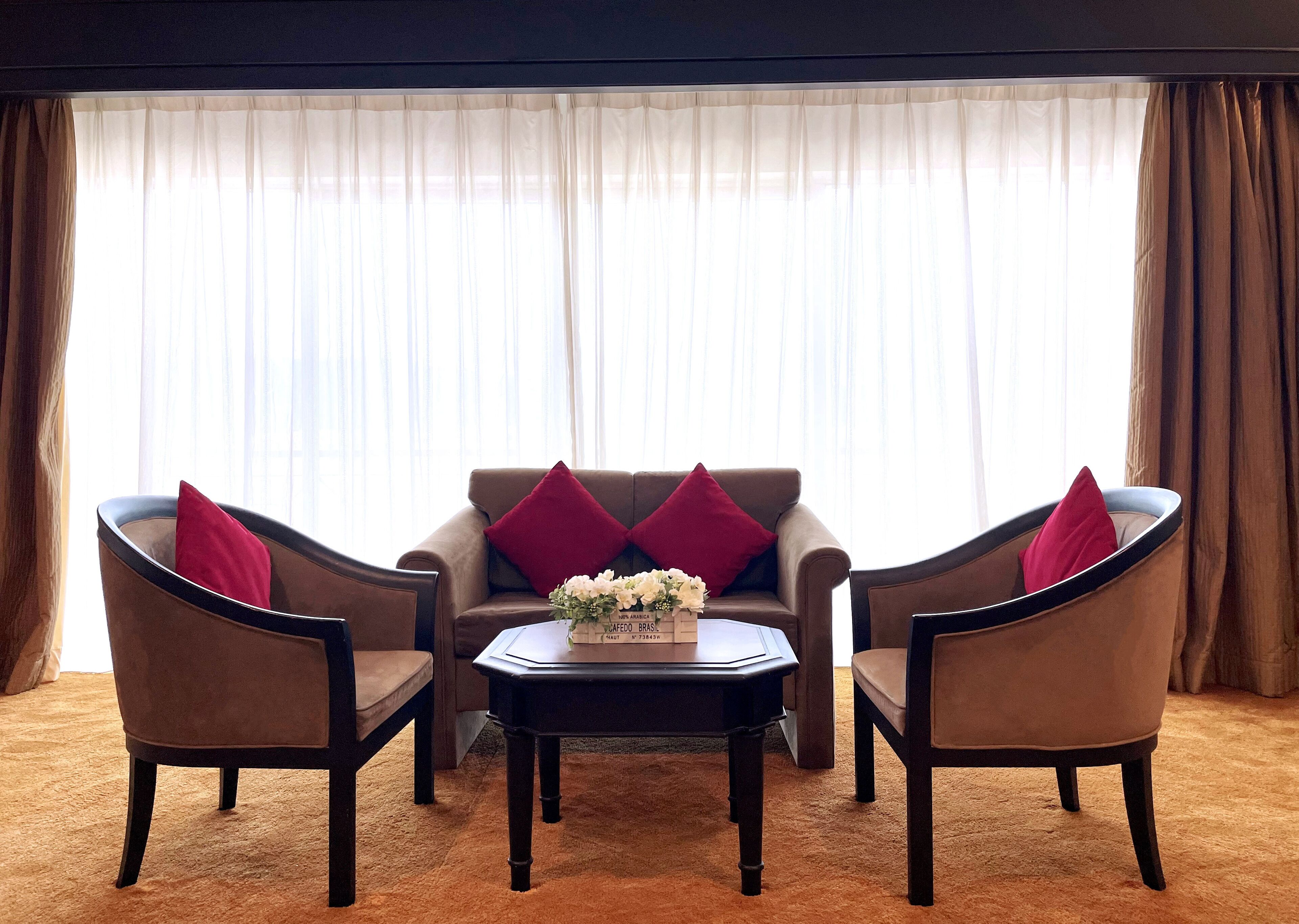 Een woonkamer met een bruine bank, twee fauteuils, een salontafel en een raam met dunne gordijnen.