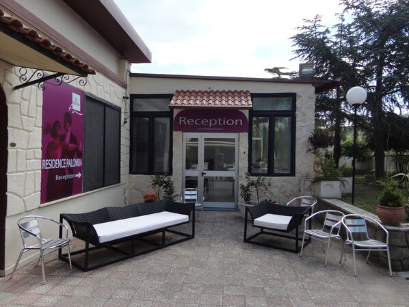 Un área de recepción con techo de azulejos, puerta de cristal y asientos exteriores con sillas de metal y un banco.