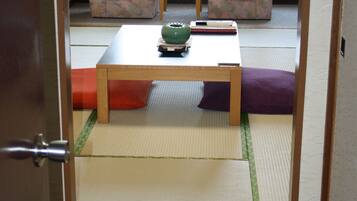 Una habitación japonesa tradicional con suelo de tatami, una mesa de madera baja, dos sillas estampadas y una ventana con cortinas.