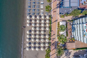 An aerial view of a beach with rows of umbrellas and lounge chairs, a palm tree-lined promenade, and a building with large windows.