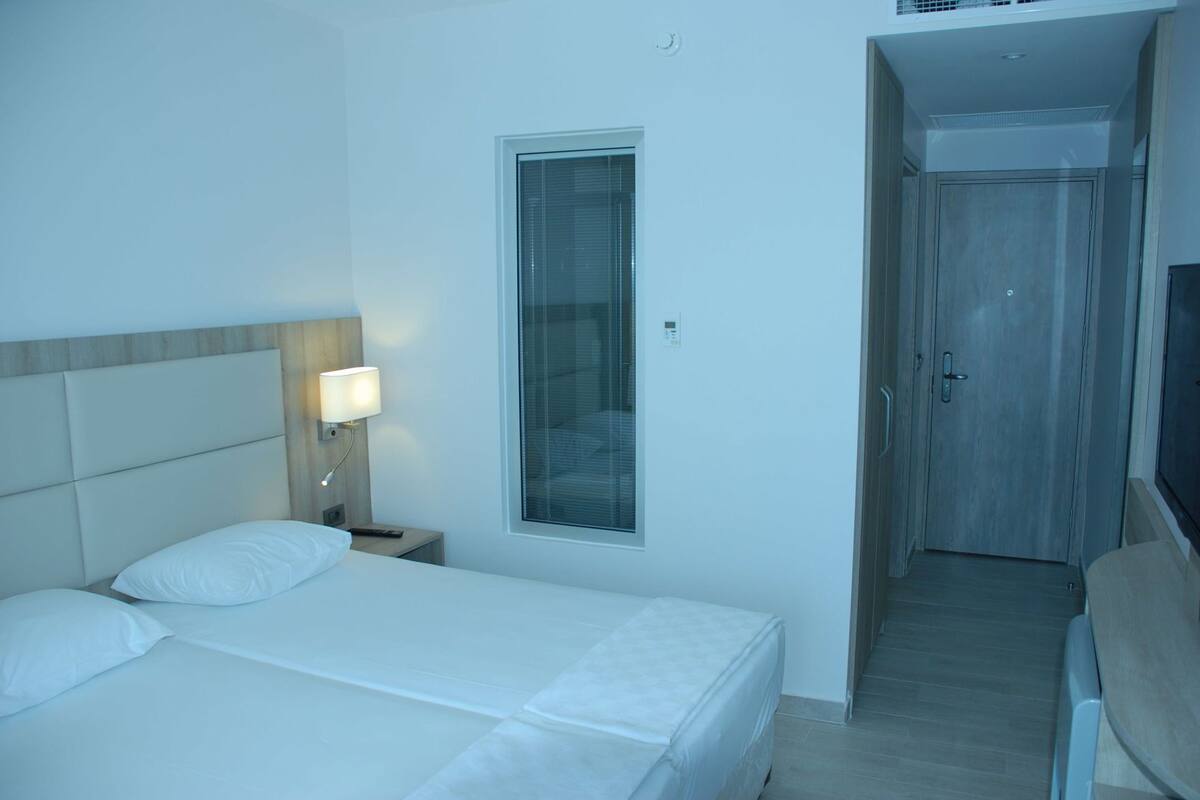 Ein Hotelzimmer mit Bett, Nachttischlampe, Fenster und einer Tür, die zu einem Flur führt.