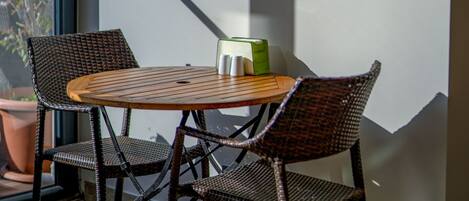 A dining area with wicker chairs and a round wooden table, a painting on the wall, and a view of trees through a window.