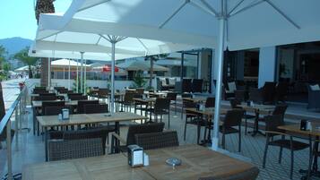 An outdoor cafe with wooden tables and chairs, white umbrellas, and a view of the street and mountains.