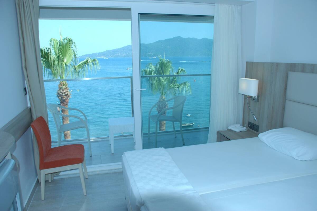 Ein Hotelzimmer mit Bett, Sessel, Tisch, Nachttisch und Blick auf das Meer und die Berge.