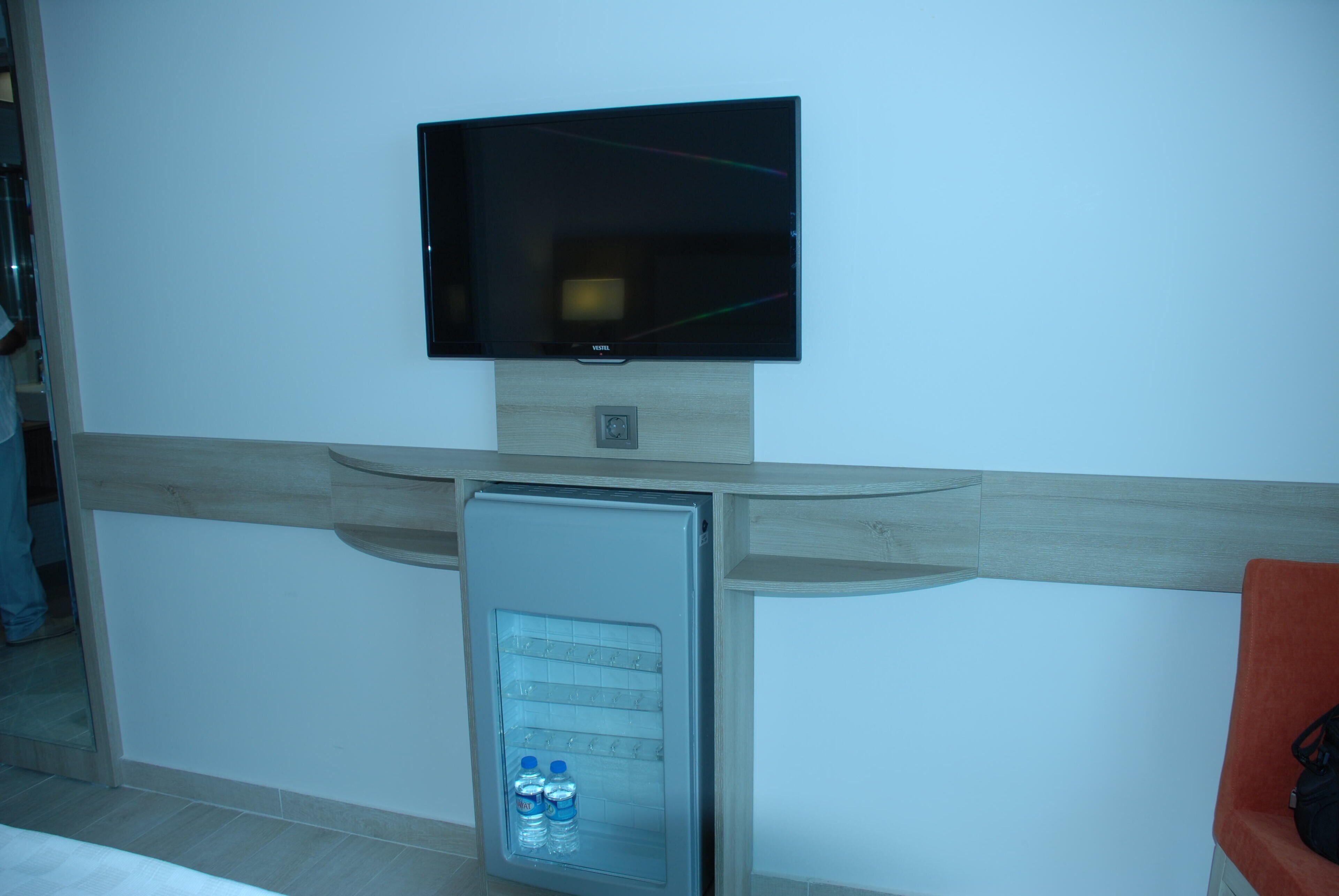 Een kamer met een wandgemonteerde flatscreen-tv, een minibar met waterflessen en een houten accentmuur.