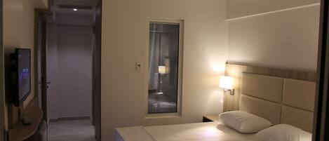 A hotel room with a bed, a TV, a window with blinds, and a nightstand with a lamp.