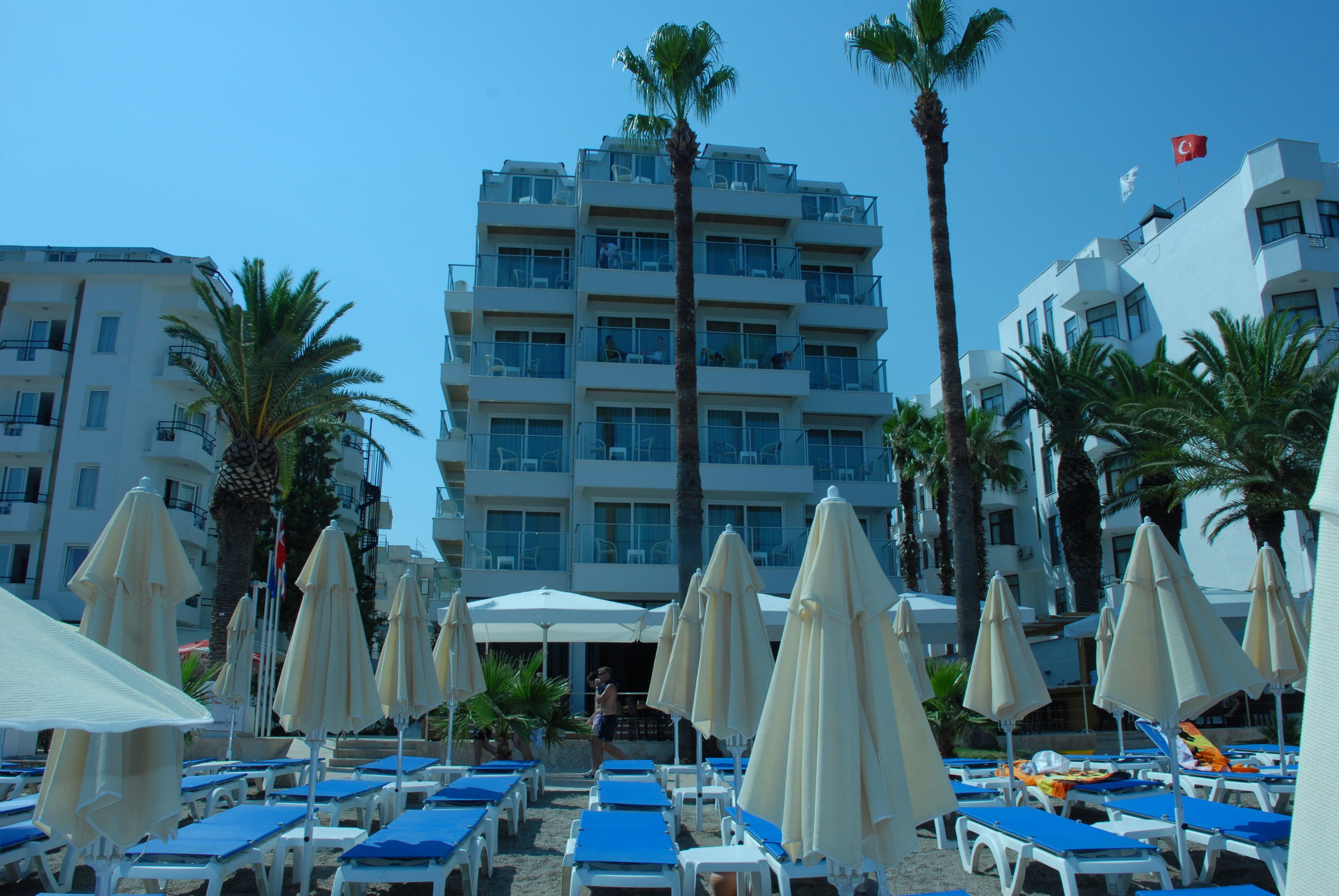 Un hotel de varios pisos, con mobiliario exterior bajo sombrillas y palmeras.