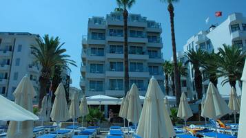 Un hotel de varios pisos, con mobiliario exterior bajo sombrillas y palmeras.
