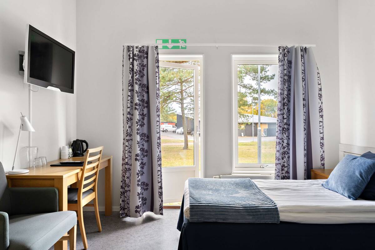 Hotelkamer met een bed, een bureau met stoel, een aan de muur bevestigde televisie en een raam met gordijnen.