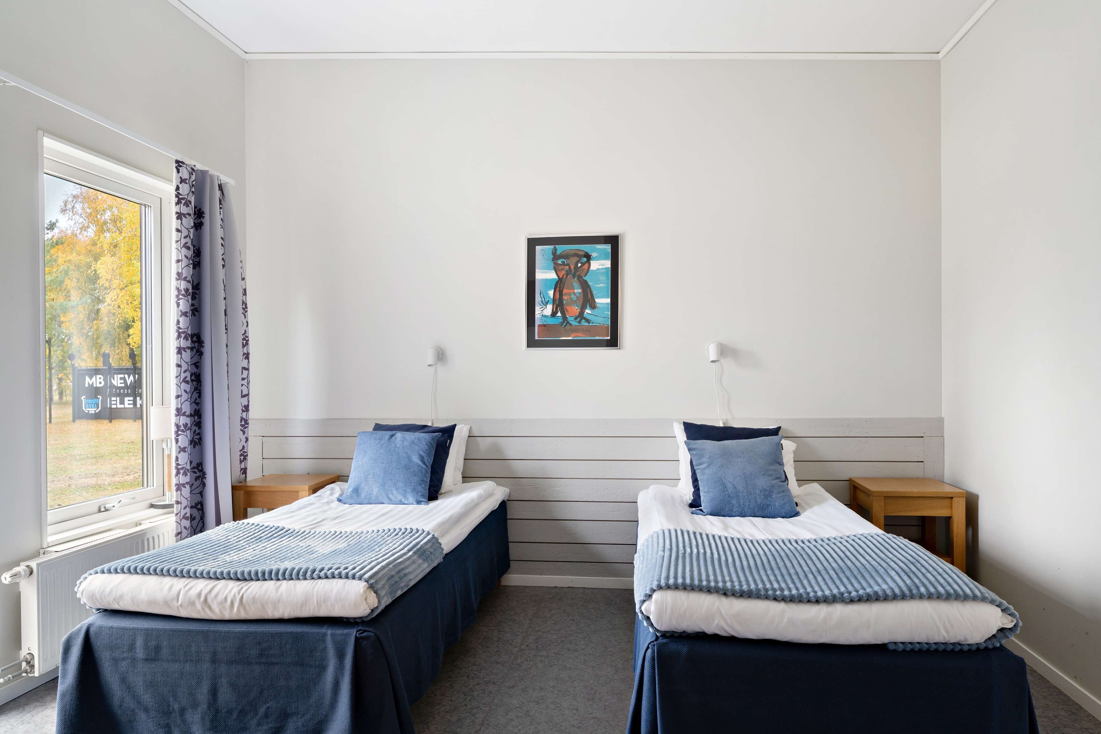 A room with two beds, a window with curtains, a radiator, a small table, and a painting on the wall.