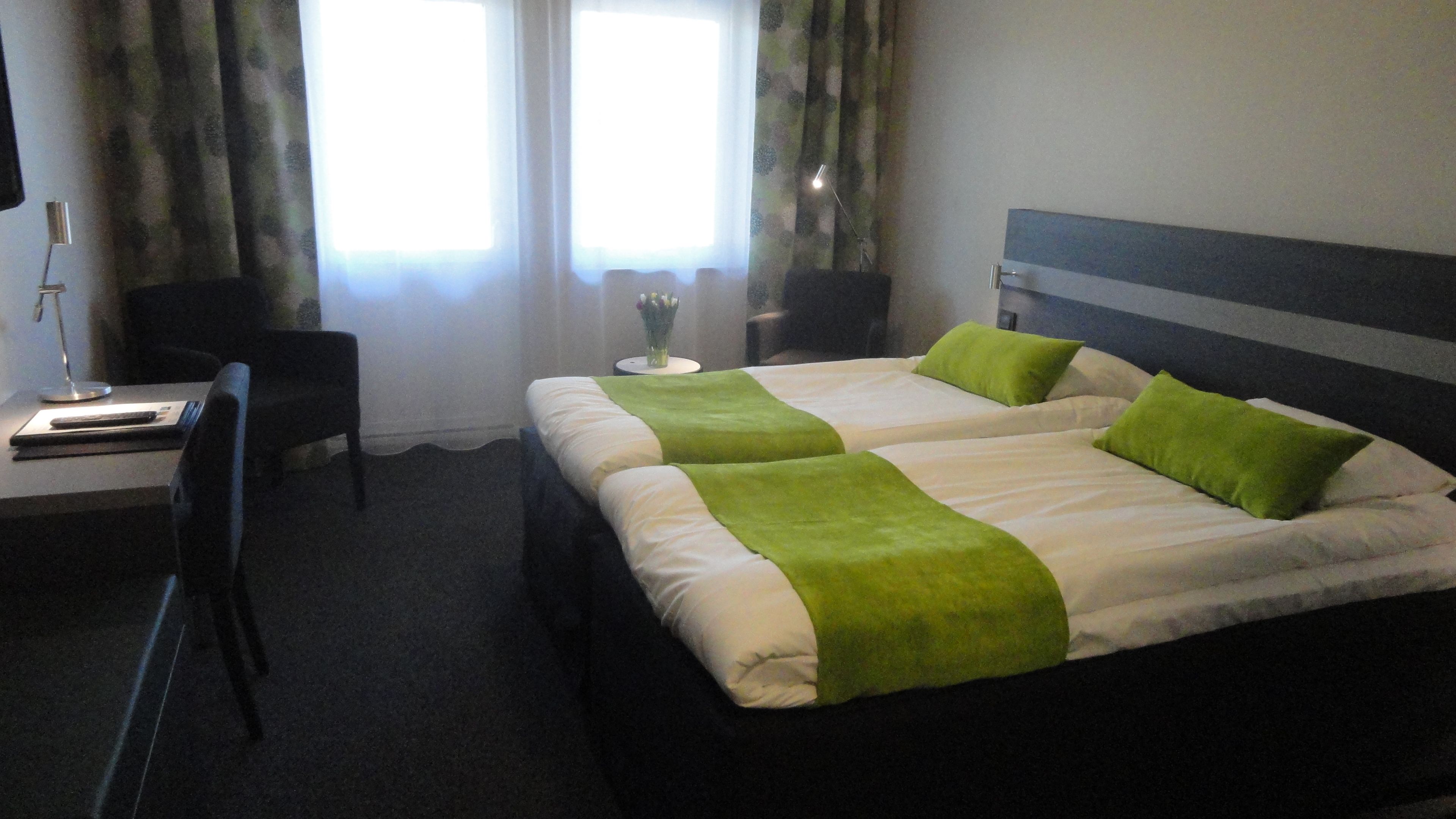 A hotel room with two beds, a desk, a chair, and a nightstand with a lamp.