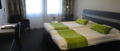 A hotel room with two beds, a desk, a chair, and a nightstand with a lamp.