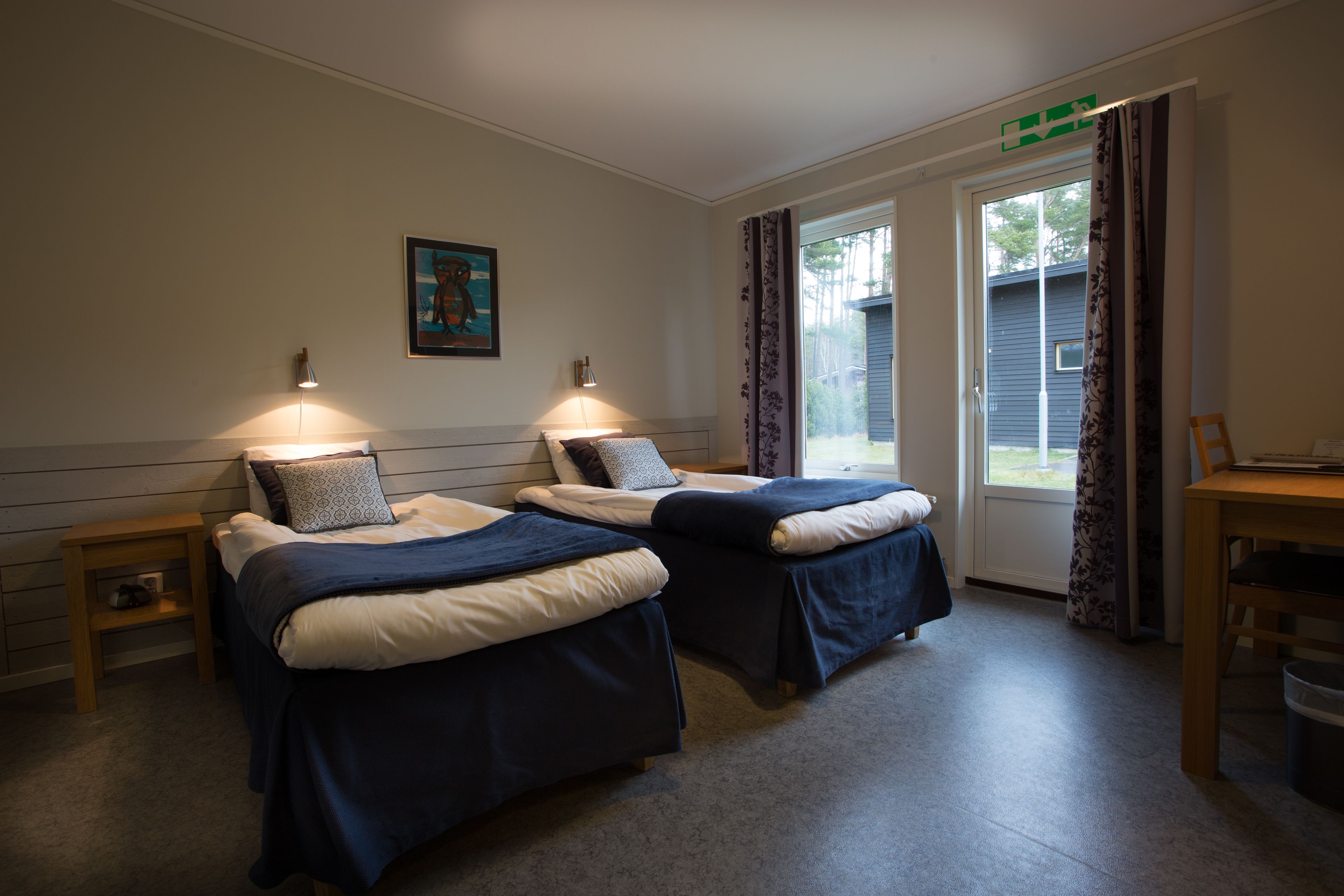 A room with two beds, a small table, and a chair. There is a painting on the wall and a window with curtains.