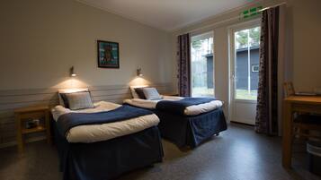 A room with two beds, a small table, and a chair. There is a painting on the wall and a window with curtains.