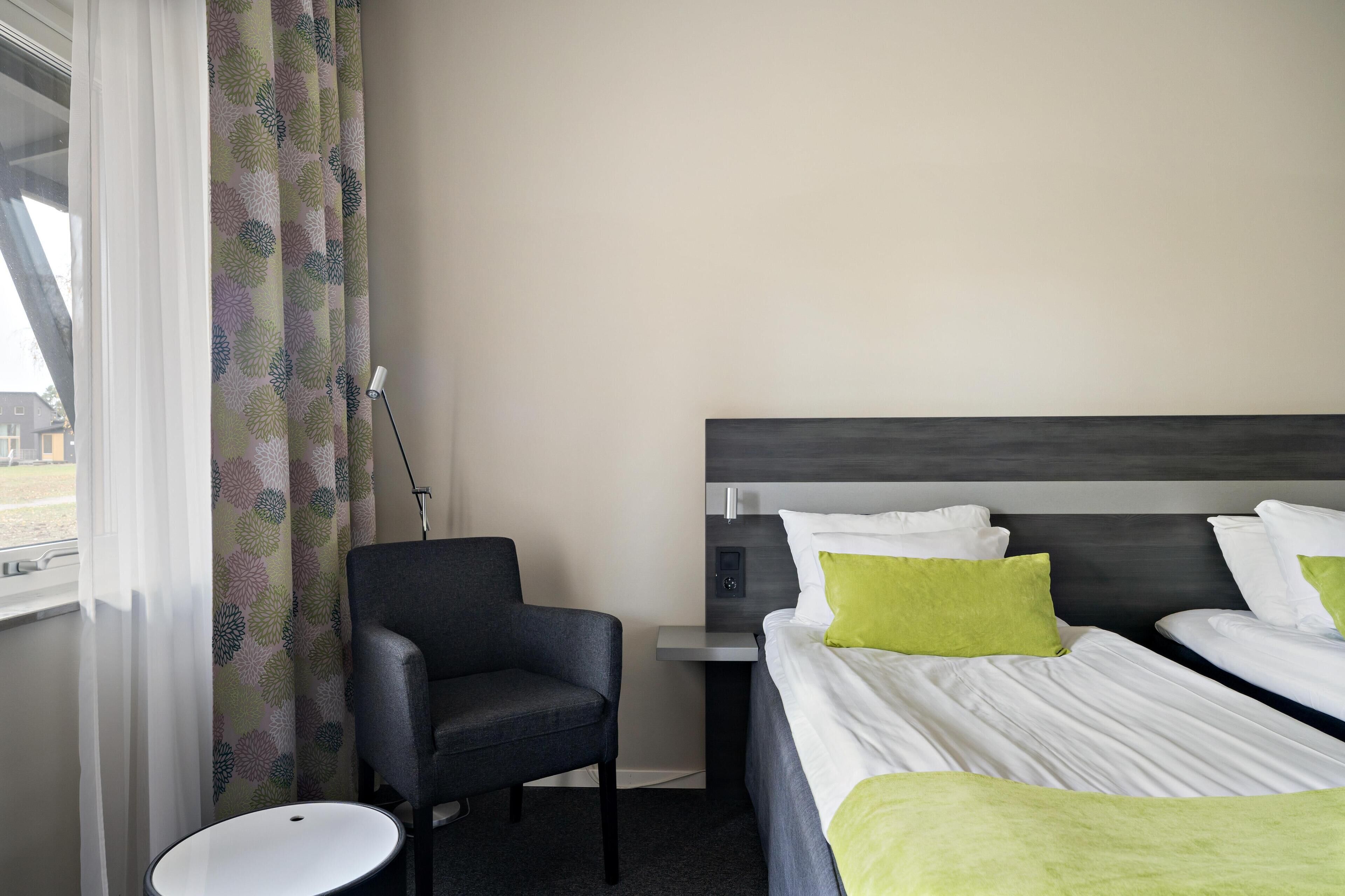 A hotel room with two beds, a chair, a bedside table, and a window with curtains.