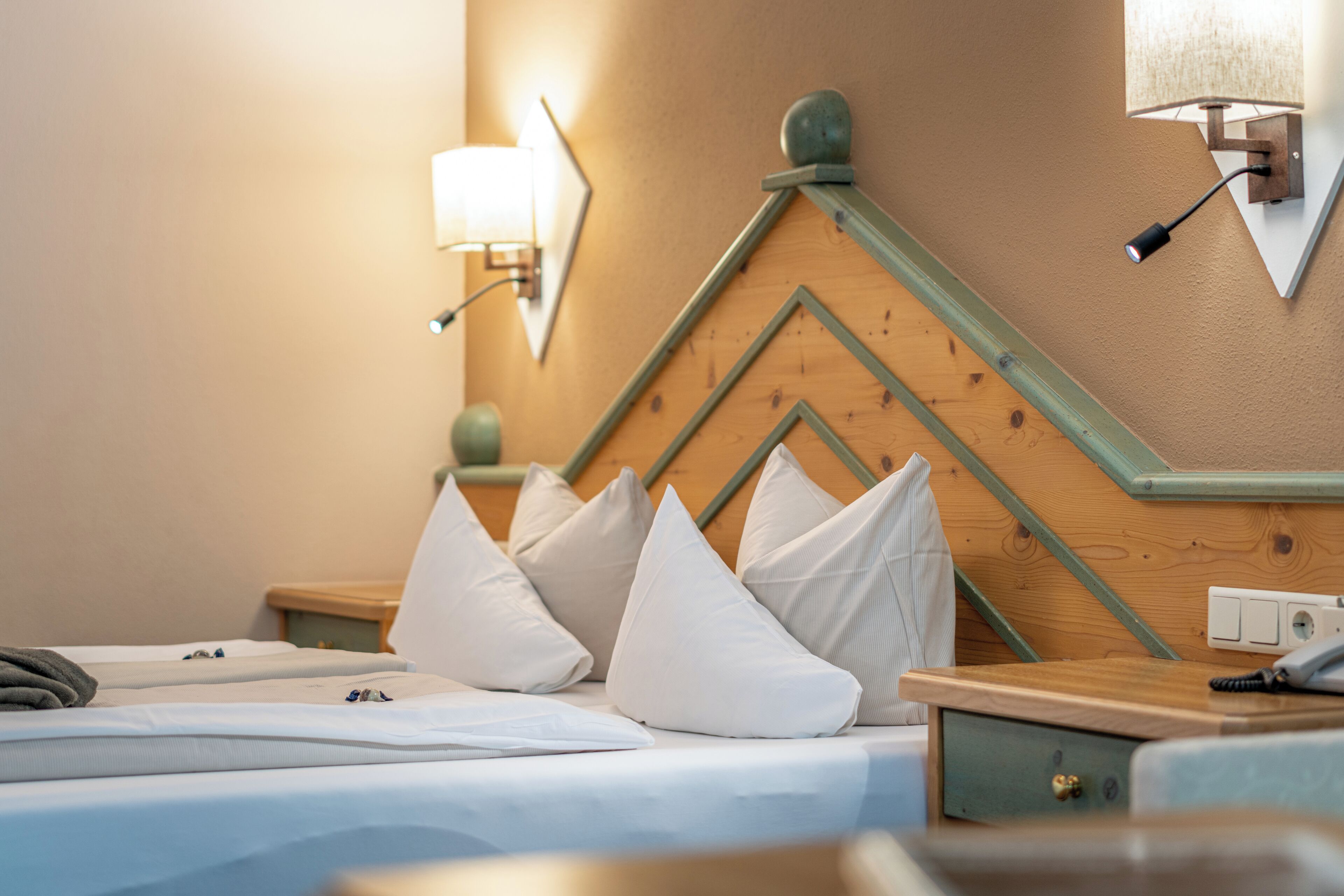 Ein ordentlich bezogenes Bett mit weißer Bettwäsche und Kissen, ein hölzernes Kopfende mit geometrischem Muster, ein Nachttisch mit Lampe sowie eine Wandleuchte.