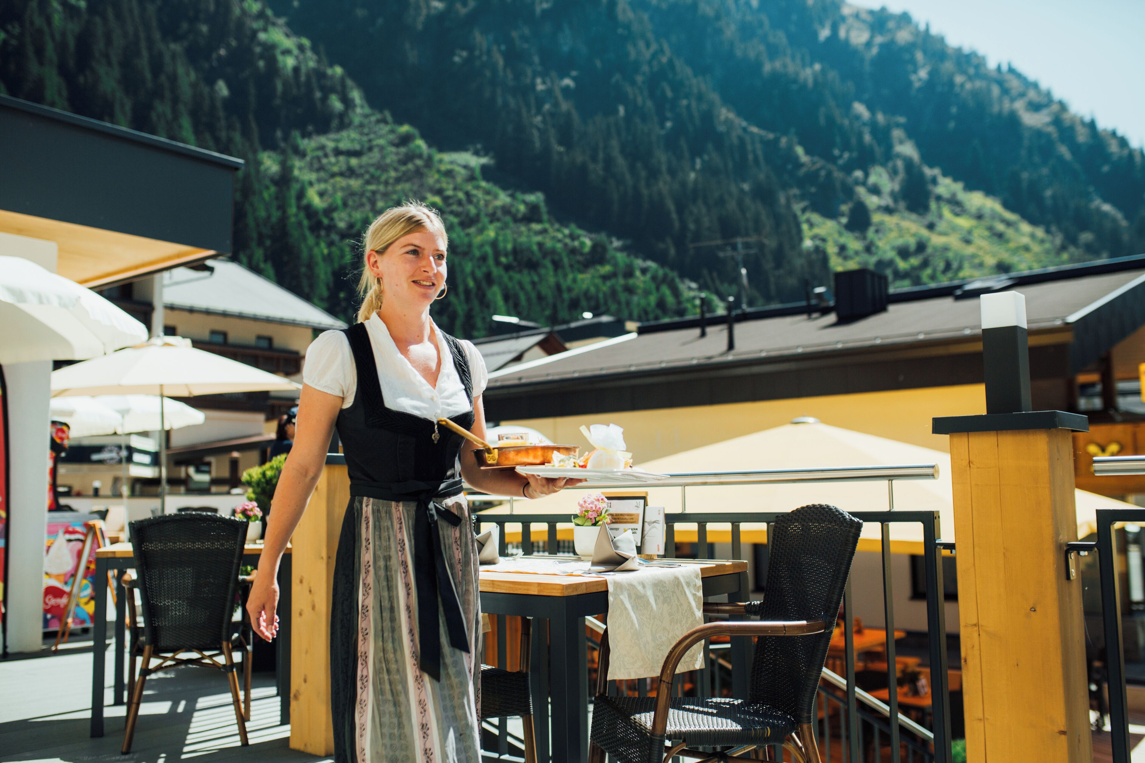 A restaurant with outdoor seating, a waitress serving food, and a mountainous backdrop.