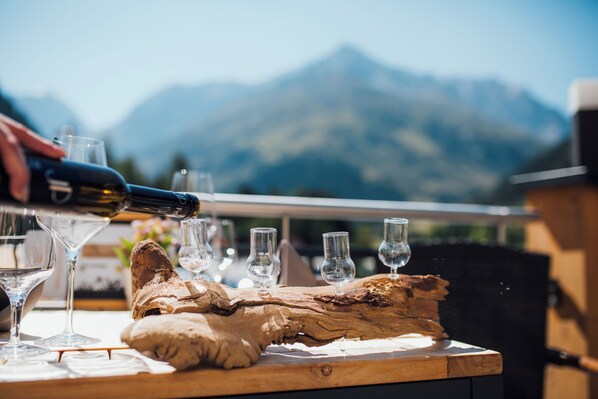 A wine bottle being poured into glasses on a wooden table with a mountain view in the background. - Hotel Mittagskogel Pitztal (Sankt Leonhard im Pitztal)