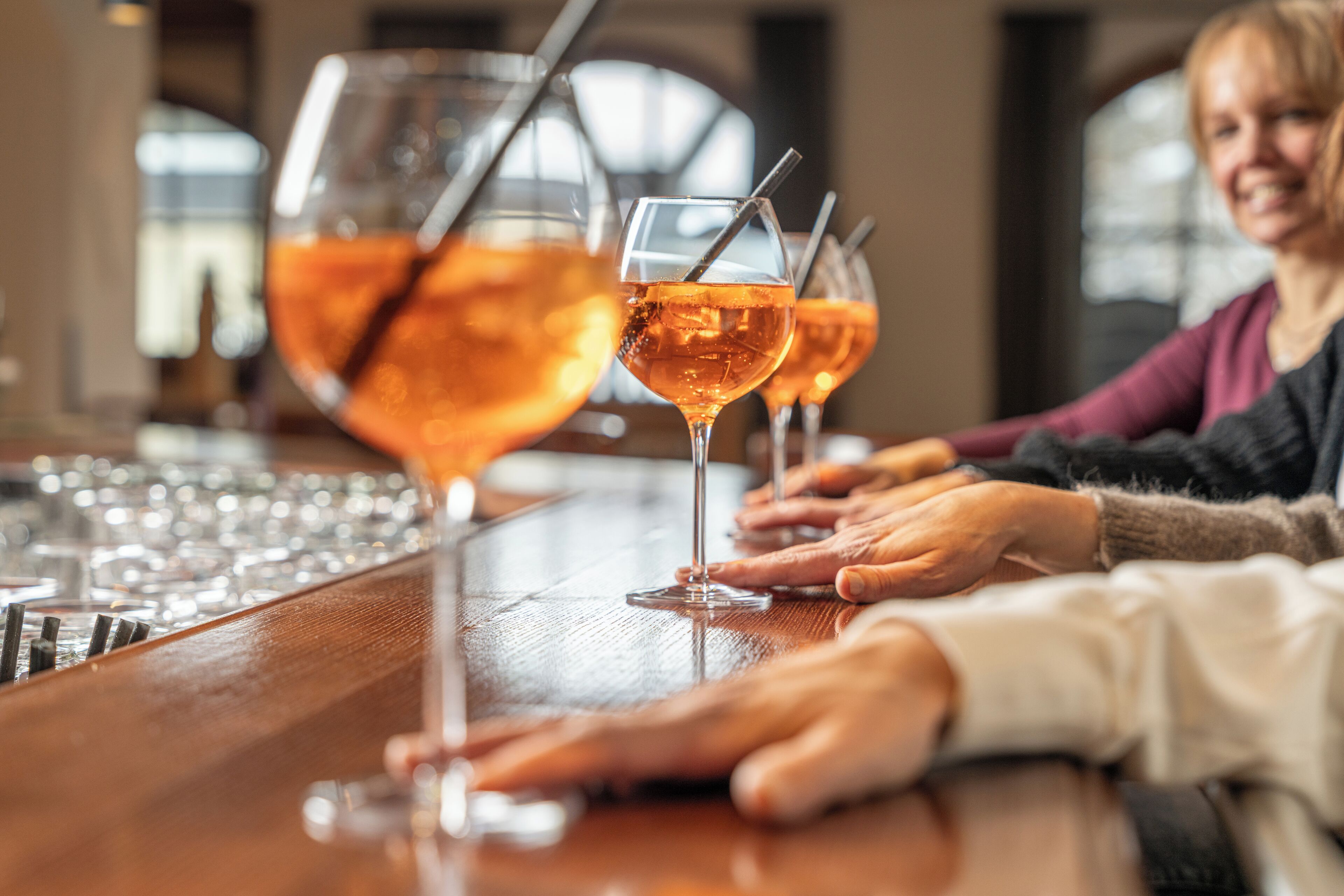 A bar setting with multiple glasses of a beverage, a bar counter, and a person’s hand resting on it.