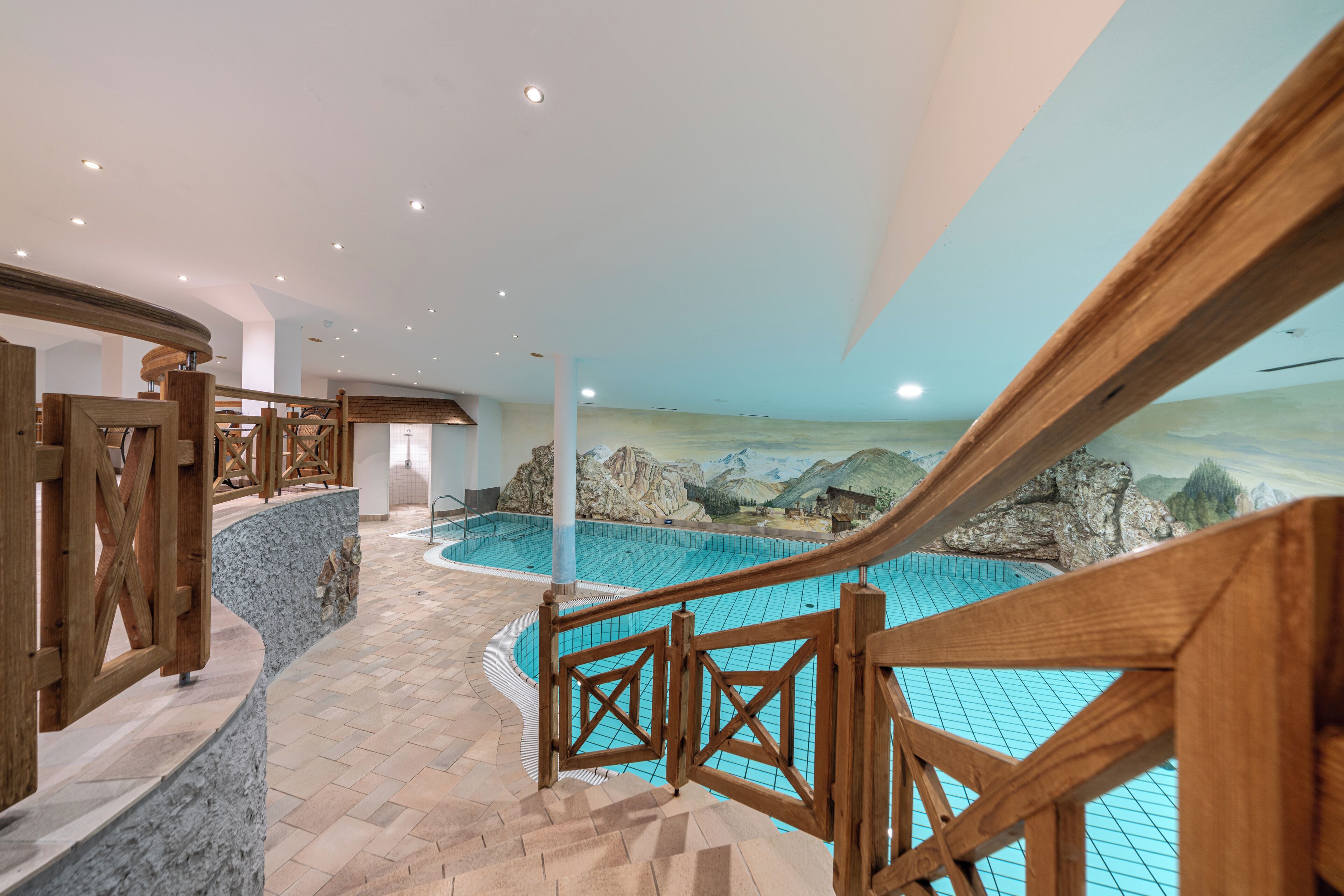 Una piscina interior moderna con una terraza de madera y un mural de un paisaje montañoso.