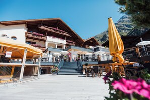 A hotel with outdoor seating, a staircase leading to the upper floor, and a mountain backdrop. - Hotel Mittagskogel Pitztal  (Sankt Leonhard im Pitztal)