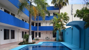 A swimming pool with a blue interior, surrounded by a building with blue and white exterior walls, palm trees, and outdoor seating. - Hotel Principe (Chetumal)