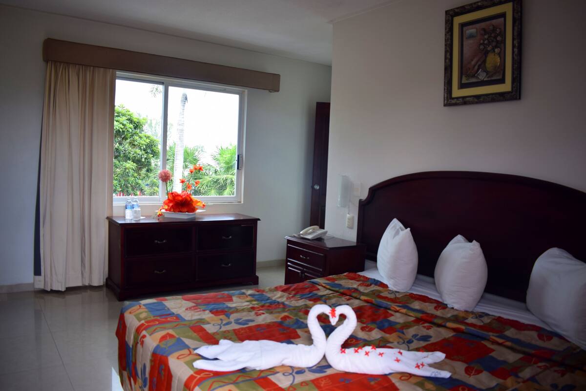 Habitación de hotel con una cama grande, un aparador, una mesita de noche, una ventana con cortinas y un cuadro en la pared.