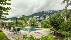 A pool area with outdoor seating and umbrellas, surrounded by lush greenery and a mountain backdrop.