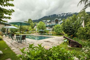 A pool area with outdoor seating and umbrellas, surrounded by lush greenery and a mountain backdrop.
