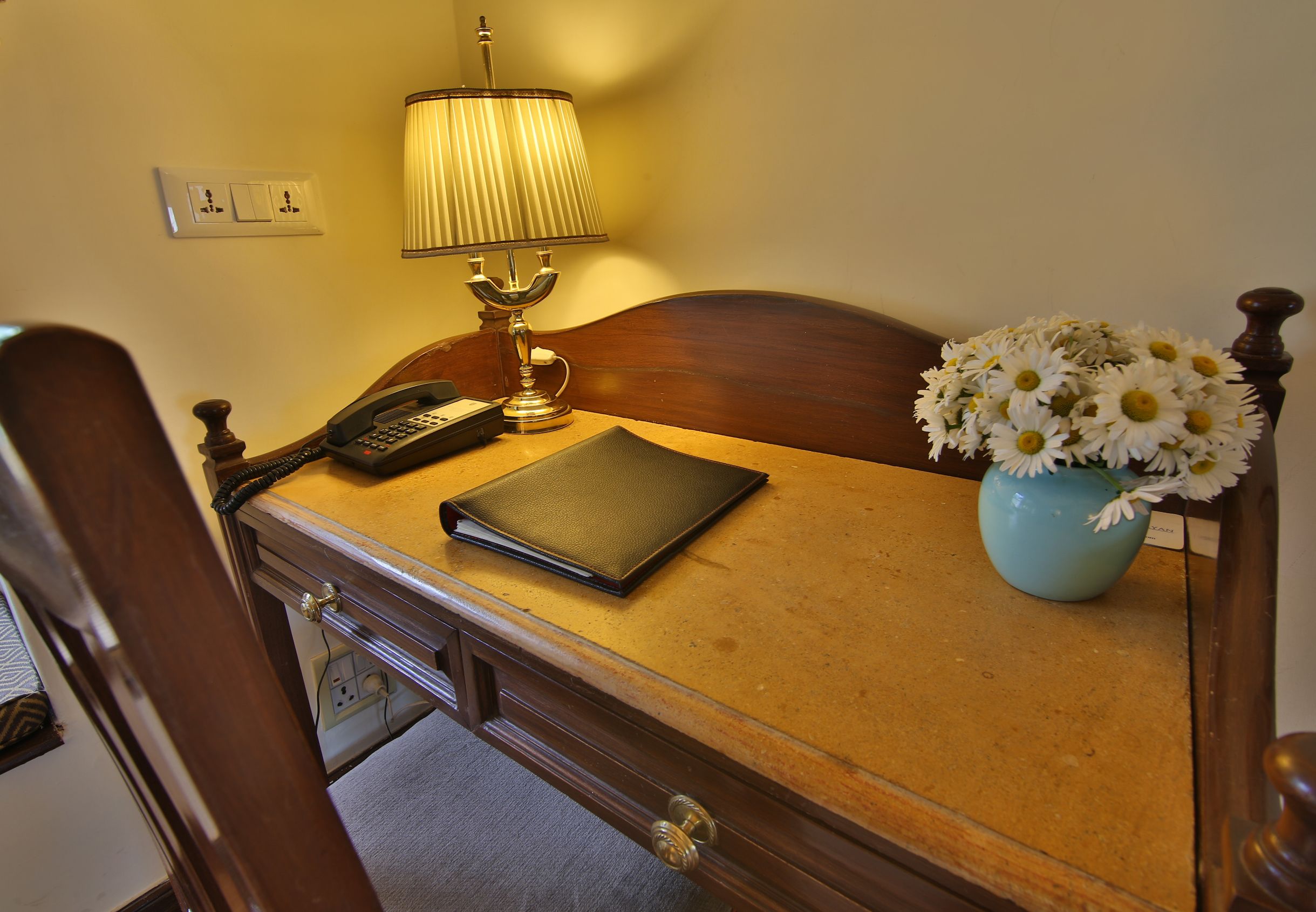 A wooden bedside table with a lamp, a phone, a notebook, and a vase of flowers.