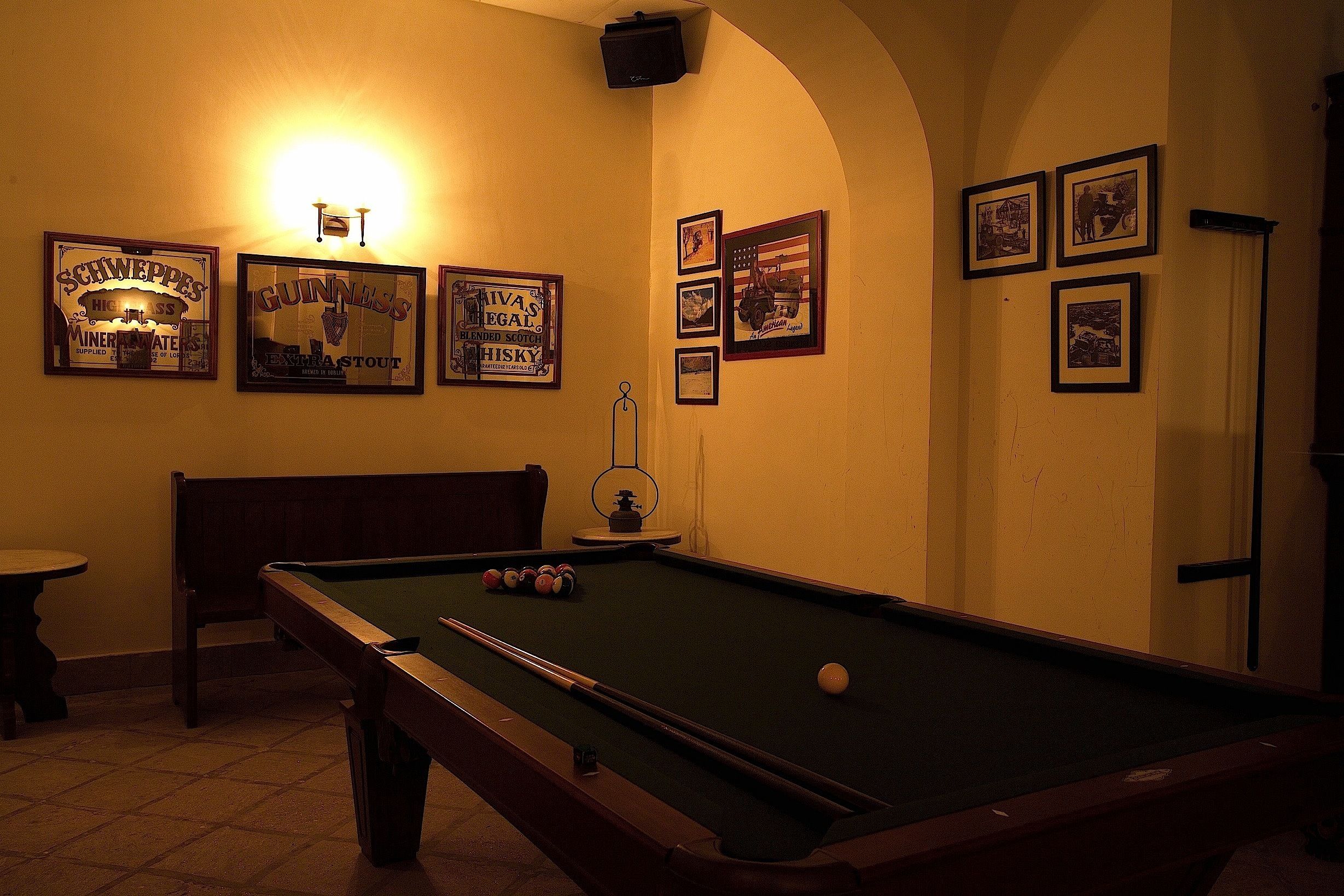 A pool table with a cue stick, a billiard ball, and a billiard rack. There are framed pictures on the wall and a small table with chairs.