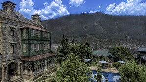 A historic stone building with green accents, a swimming pool, and a mountainous backdrop.