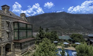 A historic stone building with green accents, a swimming pool, and a mountainous backdrop.