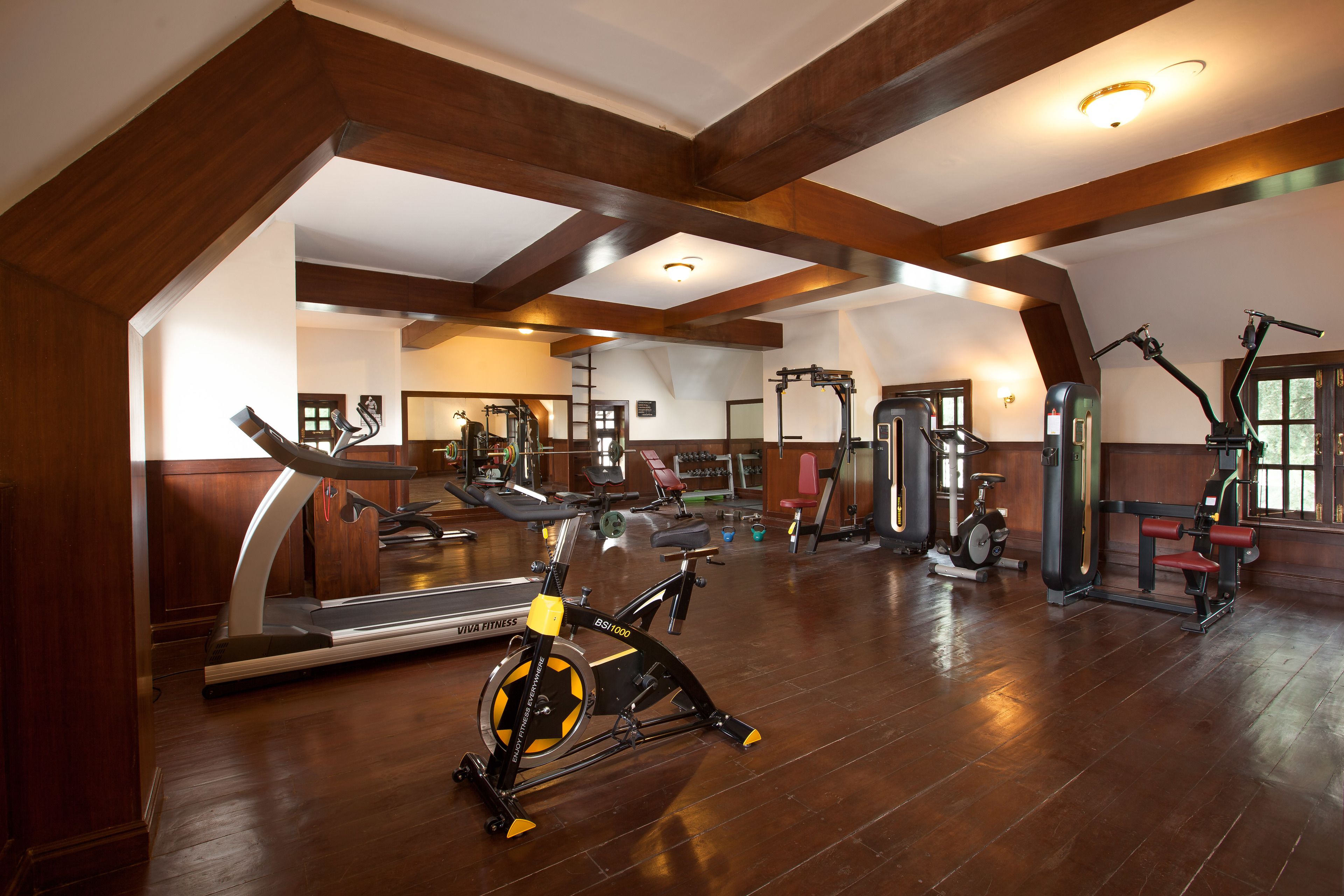 A spacious gym with various exercise equipment, wooden beams on the ceiling, and a large mirror.