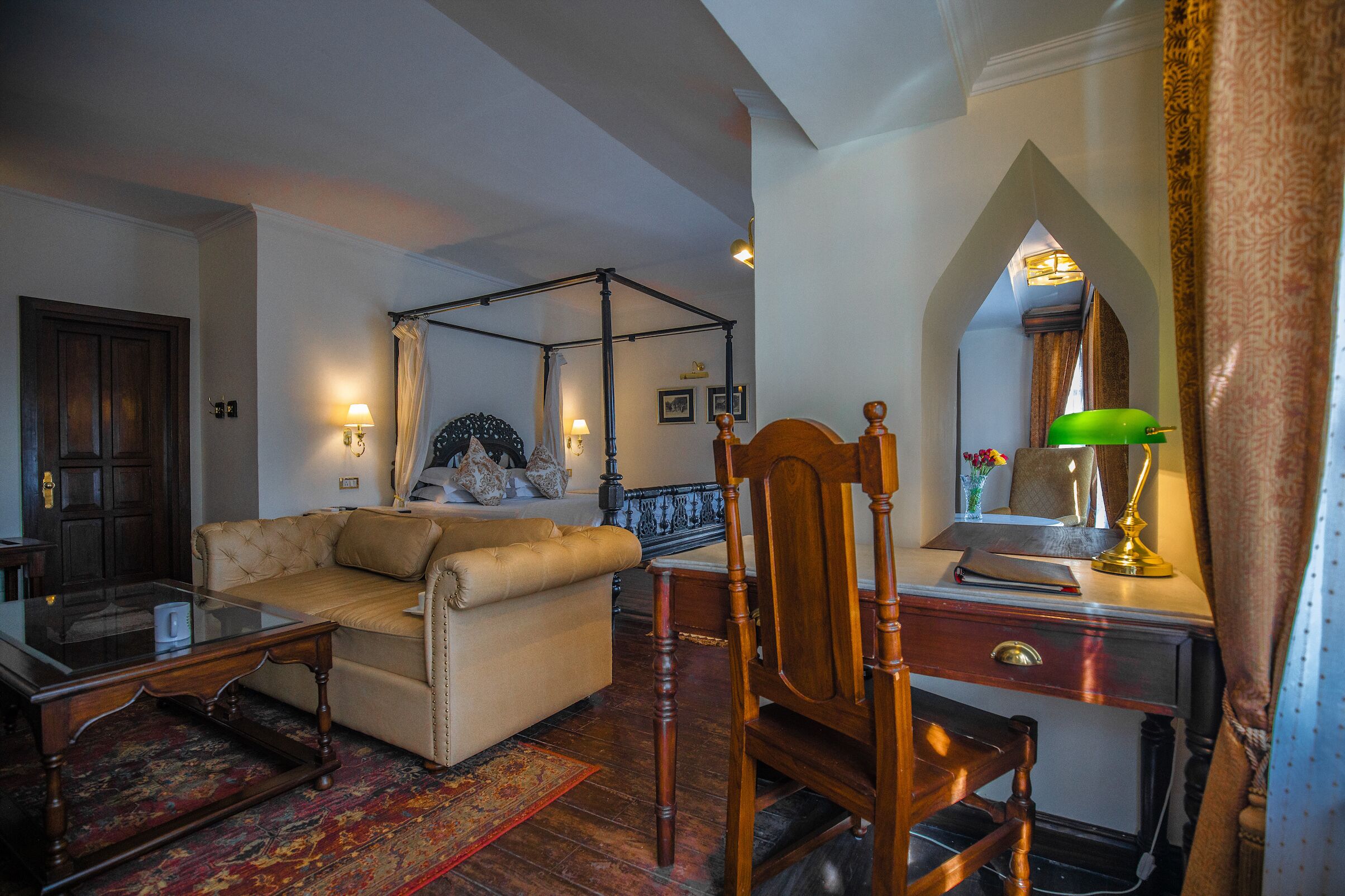 A traditional room with a four-poster bed, a wooden desk with a lamp, a leather sofa, and a patterned rug.