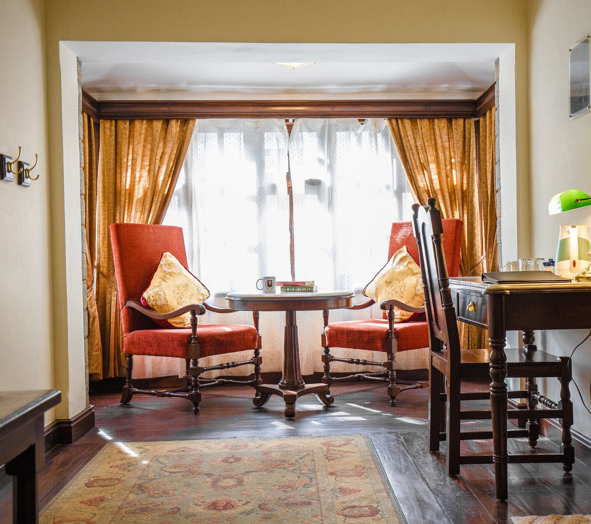 A room with a round table and two red upholstered chairs with gold cushions. There is a wooden desk with a lamp on it. The room has a large window with curtains.