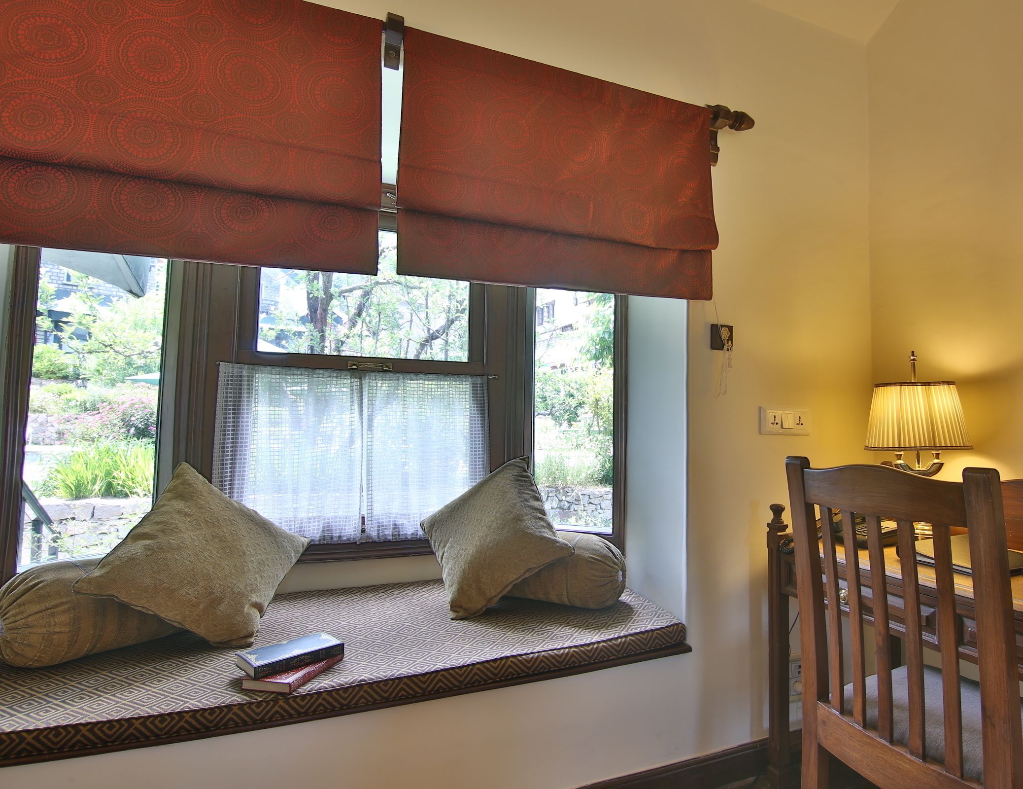 Asiento junto a la ventana con dos almohadas, un libro y una lámpara sobre una mesa.