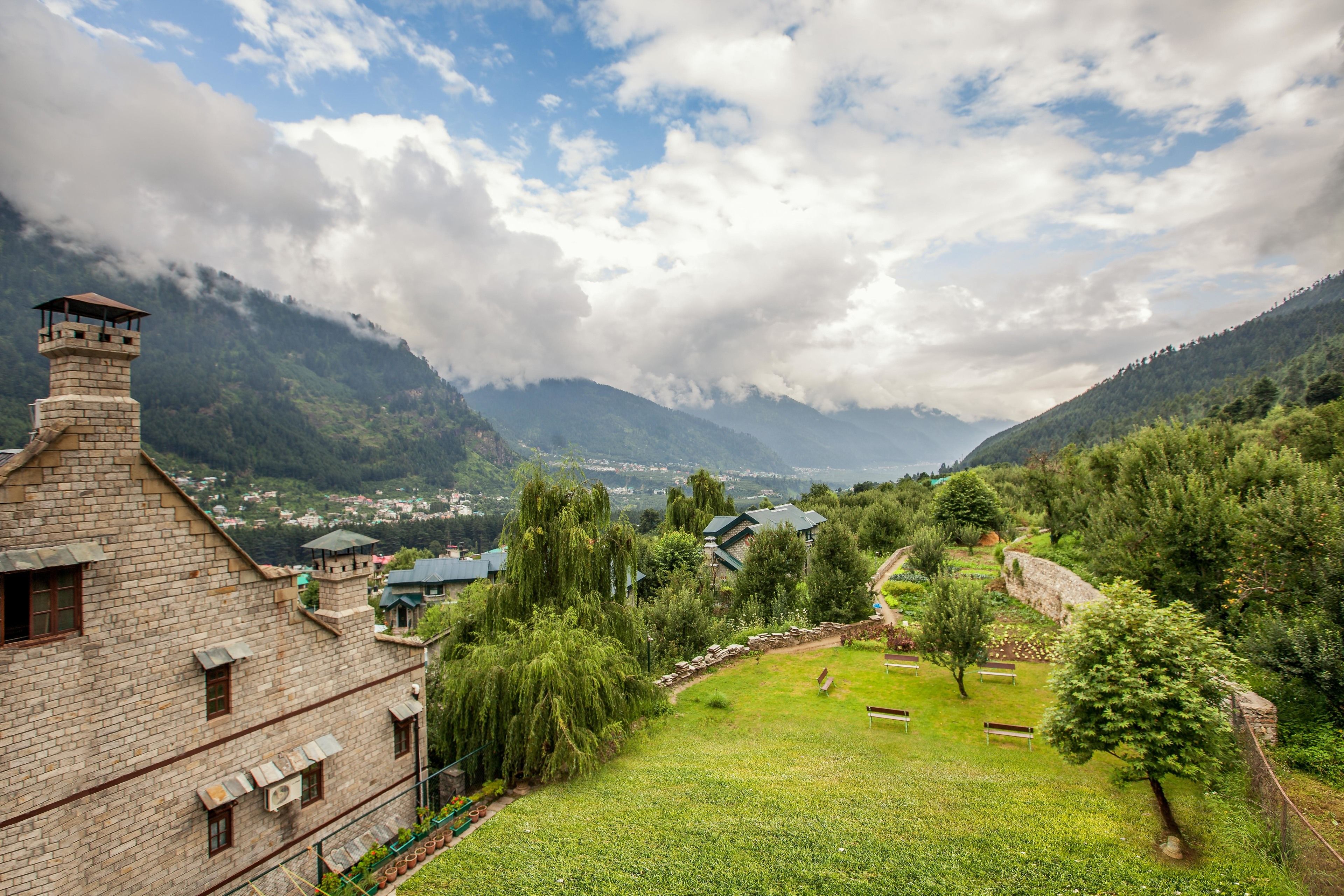 A stone building with a tower, surrounded by lush greenery and a mountainous backdrop.