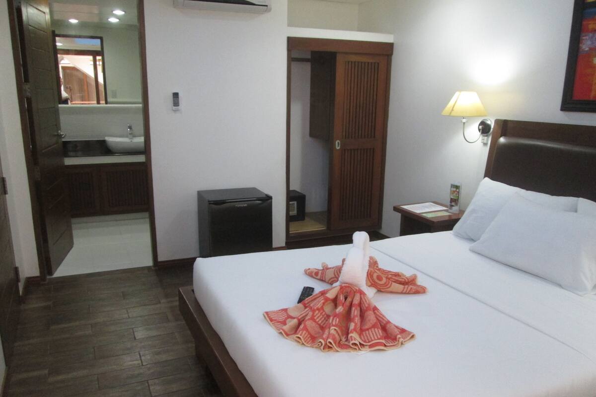 Une chambre d’hôtel comprenant un lit, une table de chevet, une lampe, un miroir, un lavabo et une porte donnant accès à la salle de bain.