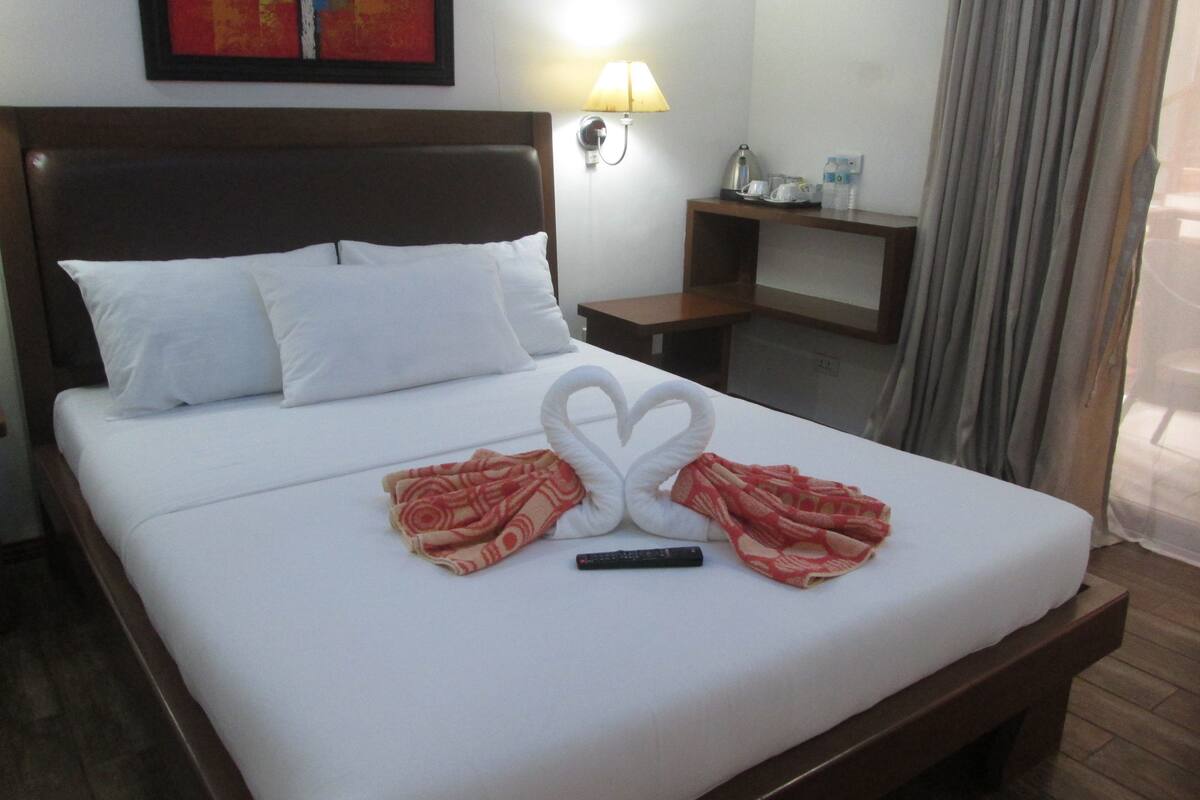Une chambre d’hôtel avec un lit bien fait, une lampe fixée au mur, une petite table avec une bouilloire et un tableau au-dessus du lit.