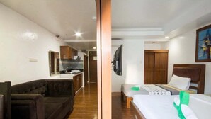Quarto de hotel com cama, sofá, televisão, escrivaninha e área de cozinha ao fundo.
