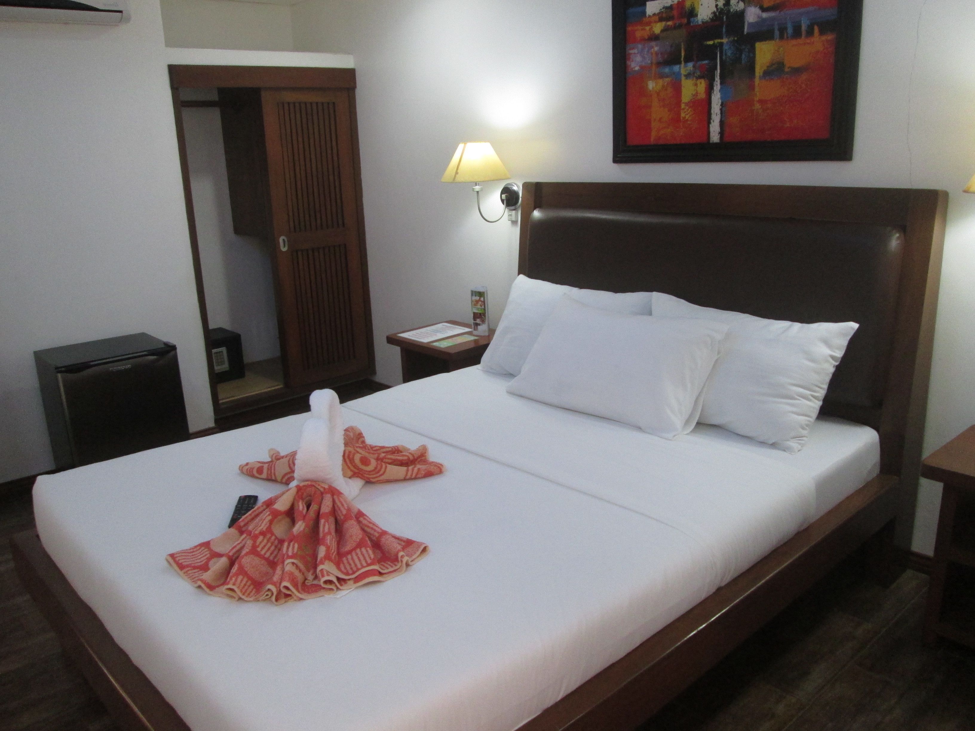 A hotel room with a neatly made bed, a wooden headboard, a nightstand with a lamp, a small trash bin, and a door leading to another room.