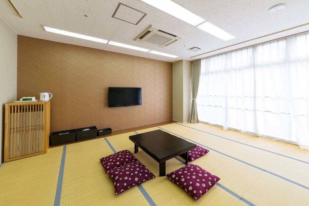 Una habitación con suelo de tatami, un televisor de pantalla plana montado en la pared, una mesa con cojines y un armario de madera con hervidor.