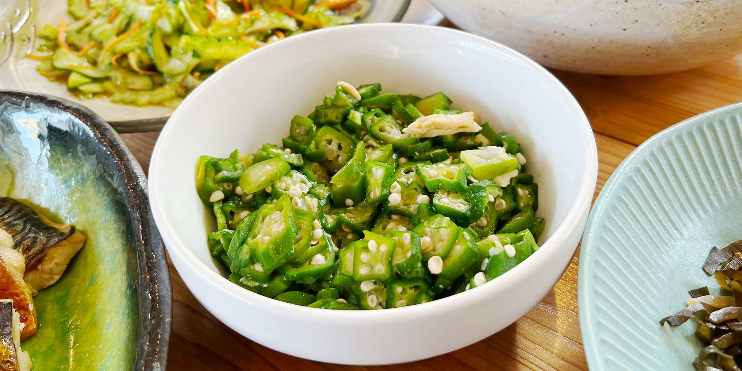 A bowl of sliced green vegetables with sesame seeds, a plate with a similar dish, and a bowl of fish on a wooden table.