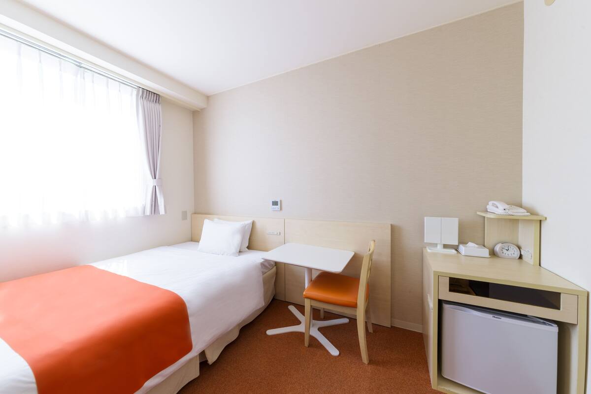 Una habitación de hotel con cama, escritorio, silla, refrigerador pequeño y mesita de noche con lámpara y teléfono.