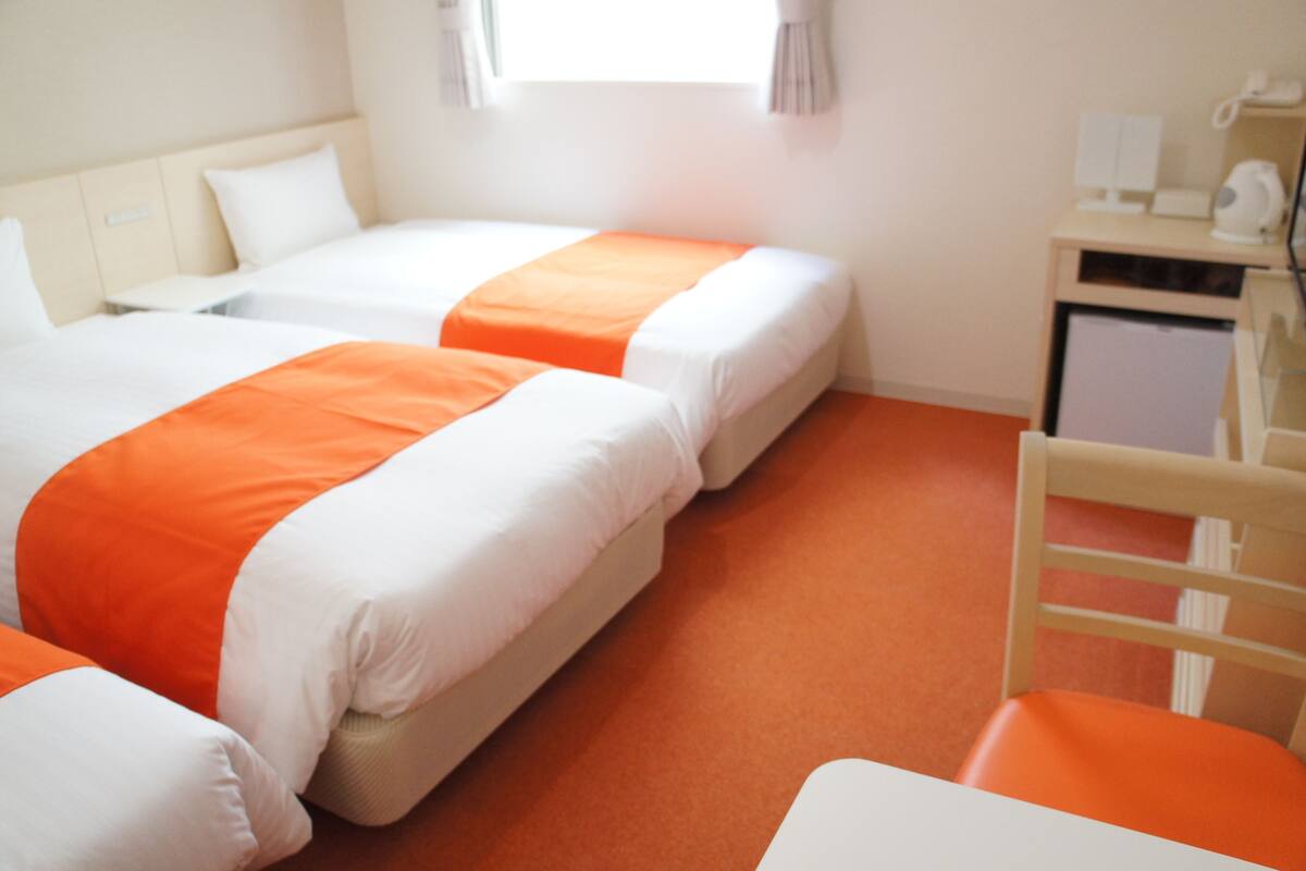 Habitación de hotel con dos camas, cada una con ropa de cama blanca y naranja, una silla de madera y un escritorio con teléfono y lámpara.